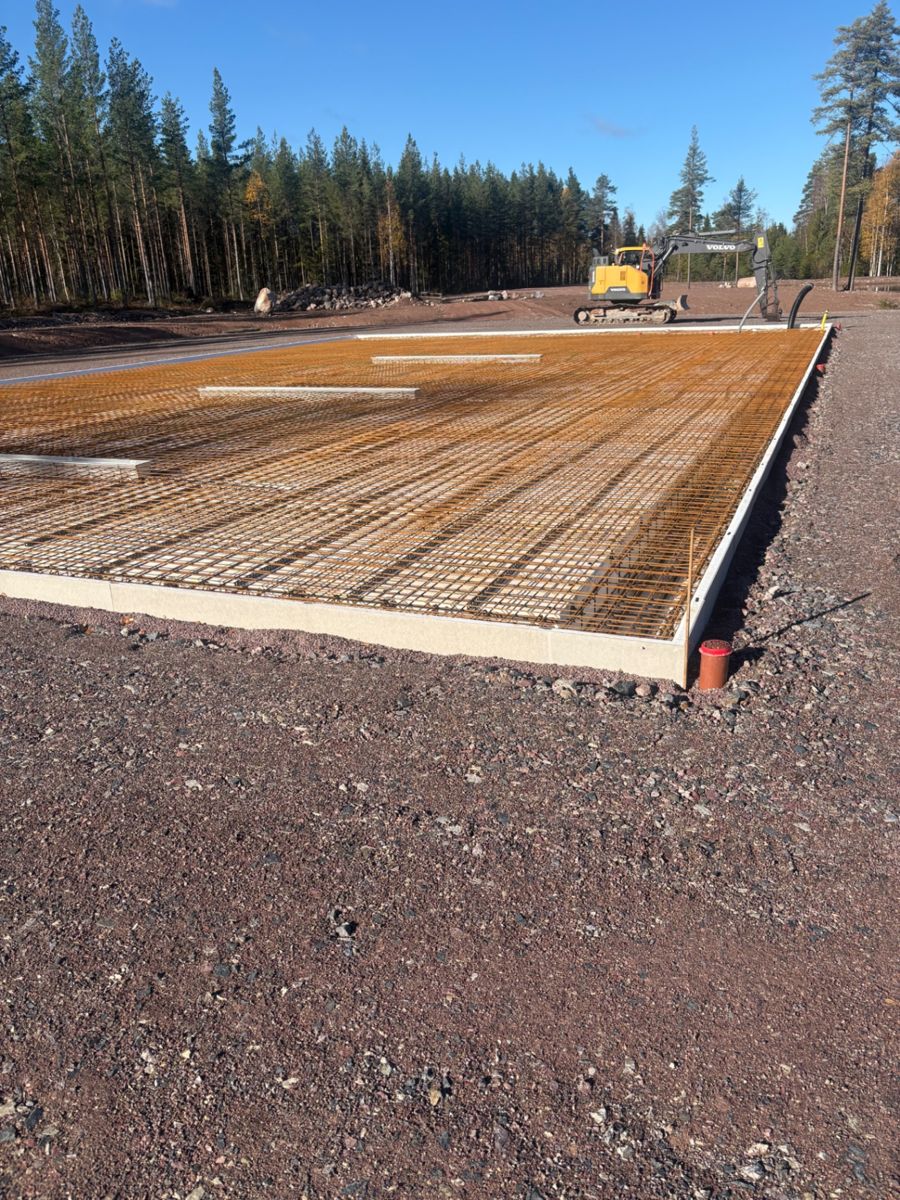 Formsatt grundbädd med utlagda armeringsnät och gul grävmaskin intill tallskog