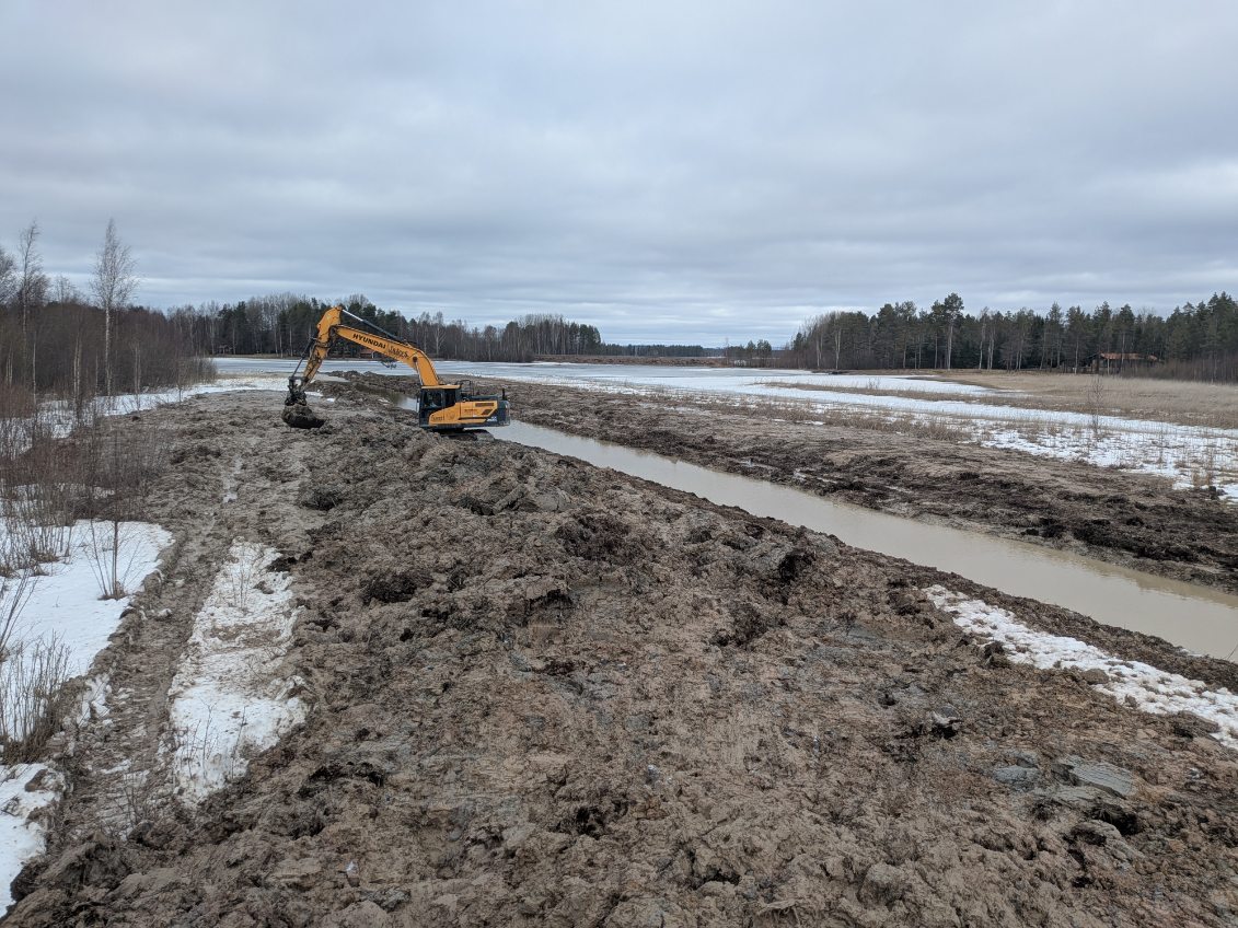Översikt över ett långt vattenfyllt dike med högar av uppgrävda sediment och en grävmaskin som arbetar längs sträckan